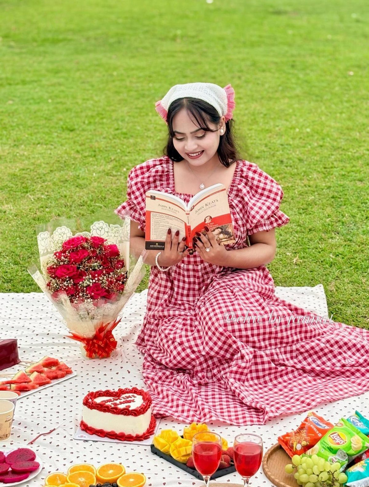 A woman in a red gingham dress reads a book during a picnic on a grassy lawn.