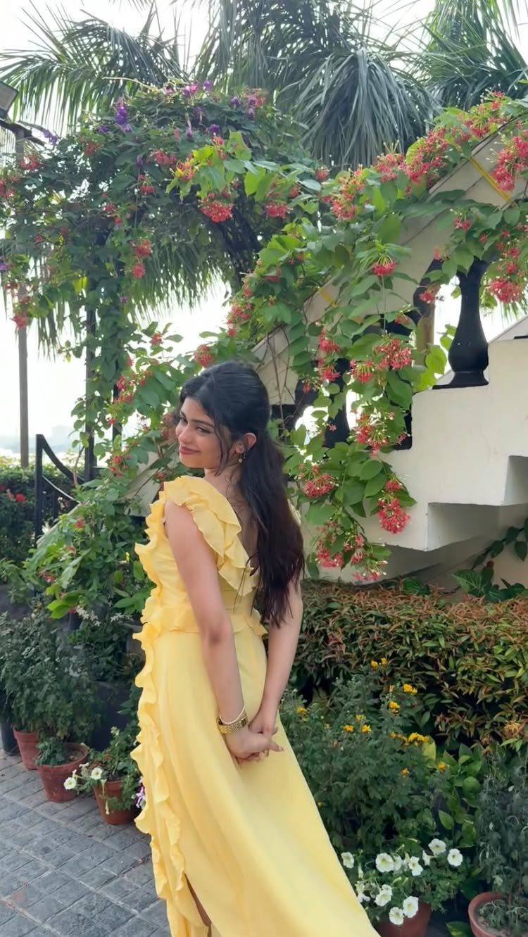 A woman in a ruffled yellow maxi dress standing amidst lush green garden plants.