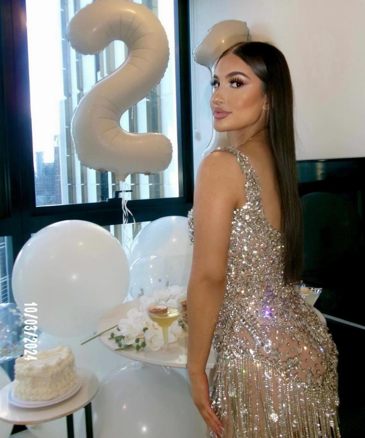 Woman in a sparkling silver sequined mini dress posing near white balloons and a cake.