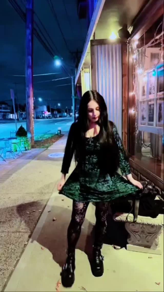 A woman wearing a dark green velvet mini dress and black lace tights outdoors at night.