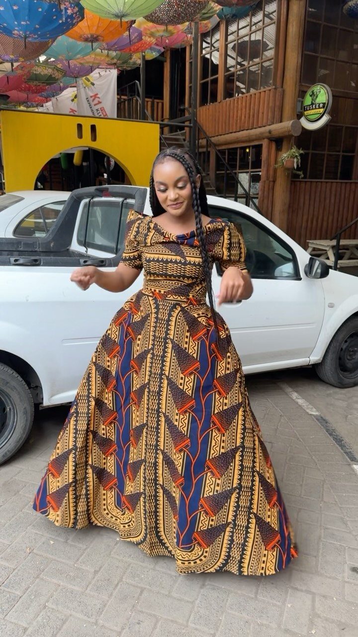 A woman wearing a patterned African print maxi dress standing in front of a white car.