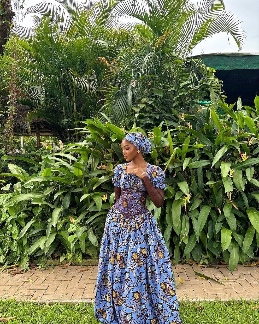 Woman in a blue patterned African print dress with flared skirt standing in front of tropical plants.