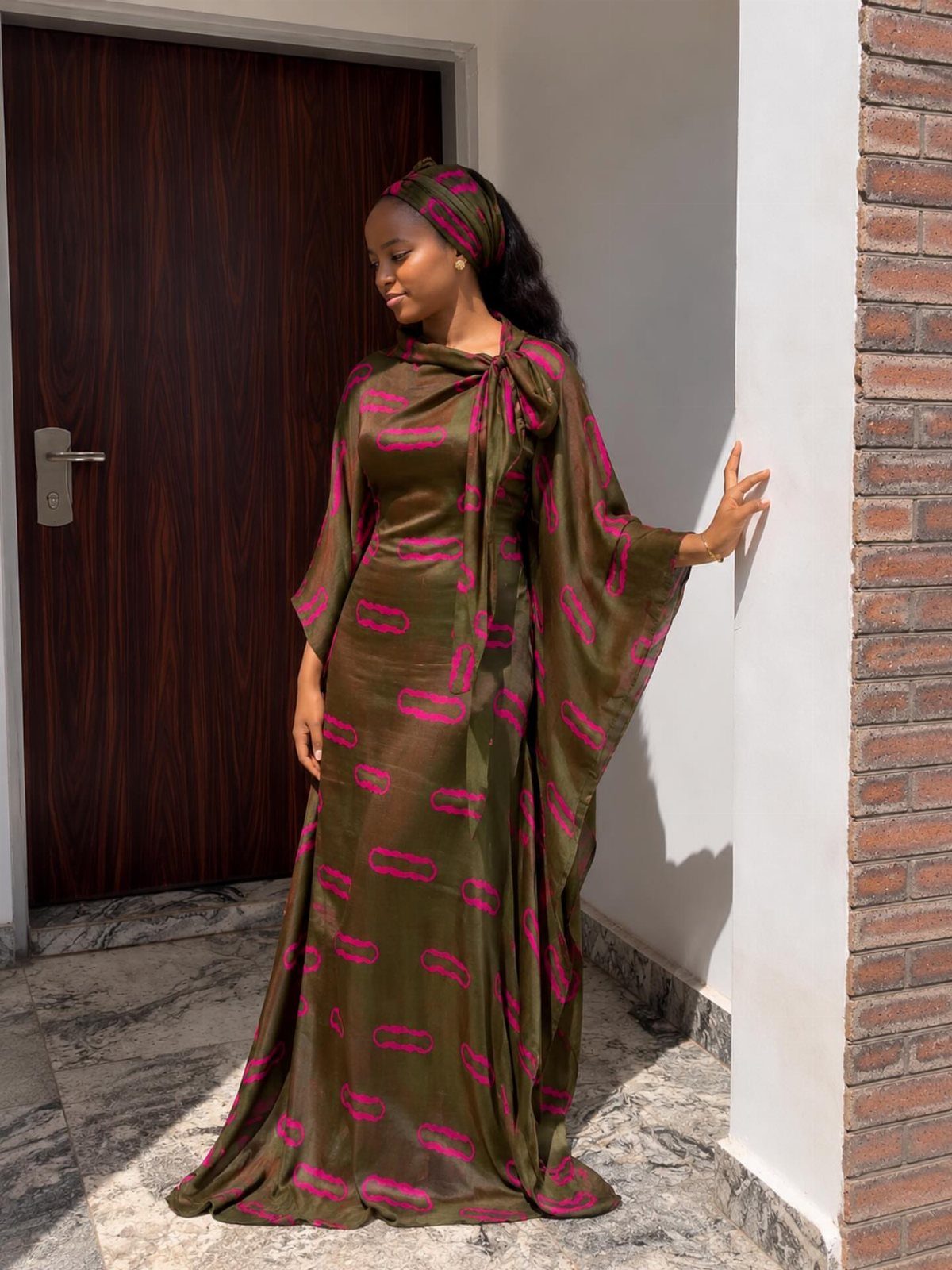 A woman wearing a voluminous olive green and pink patterned boubou and matching headwrap.