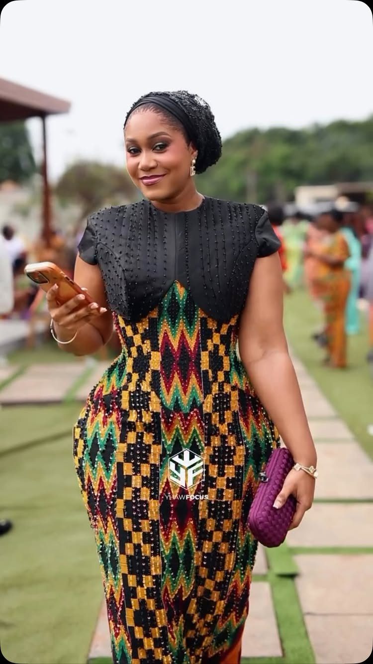 A woman wearing a colorful Kente cloth dress standing outdoors on a lawn.