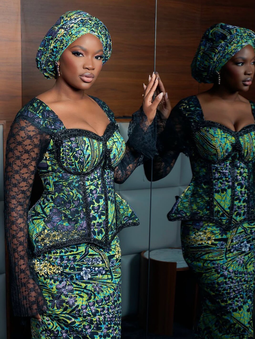 Woman in green patterned African print peplum top, matching skirt, and headwrap posing near a mirror.