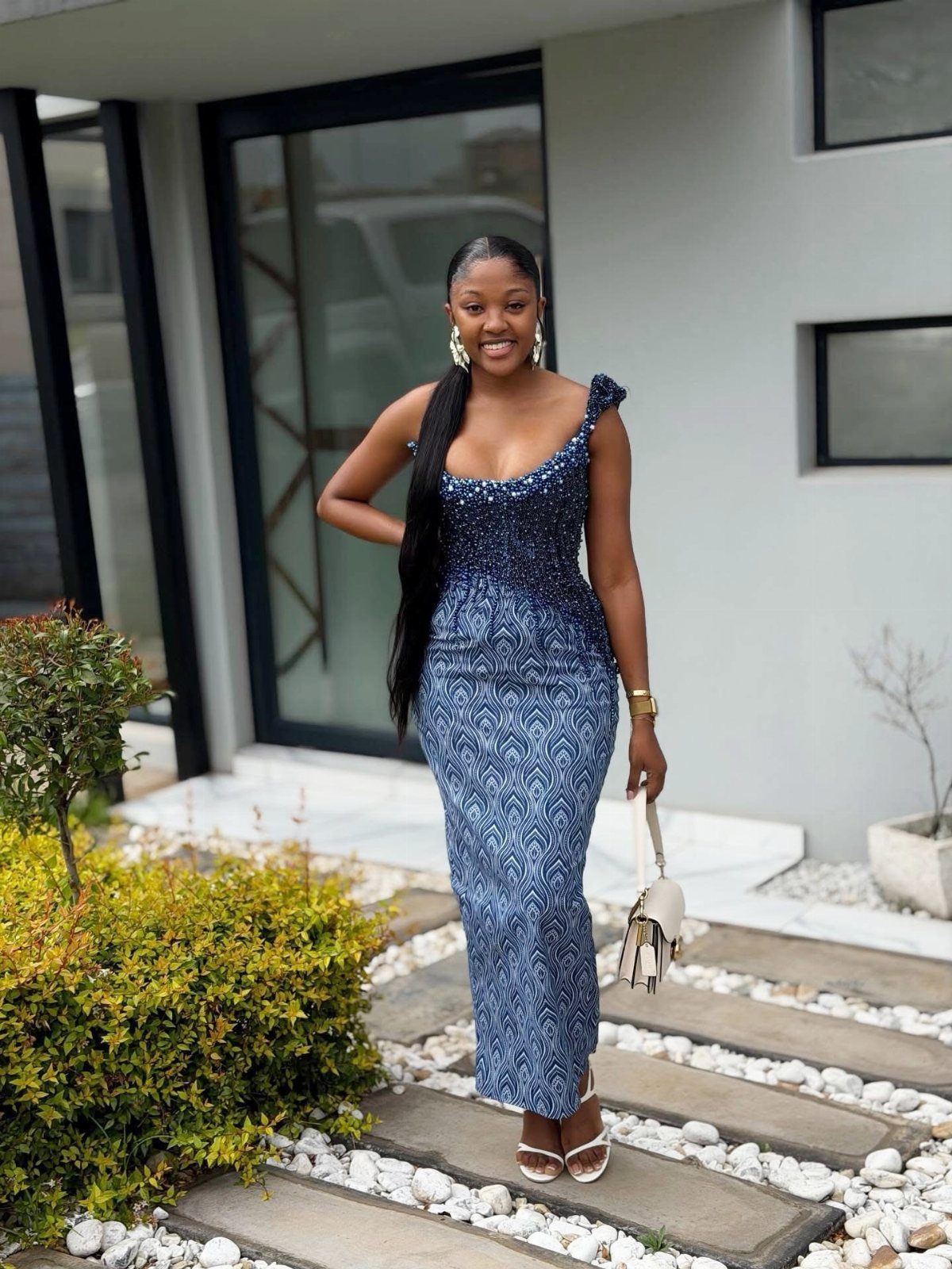 A smiling woman posing in a long, blue patterned sleeveless dress outdoors.