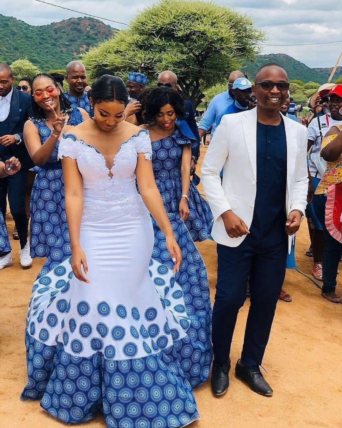A bride in a white and blue mermaid gown walks among wedding guests outdoors.