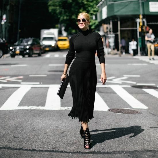 A woman in a black turtleneck midi dress walking across a city street crosswalk.
