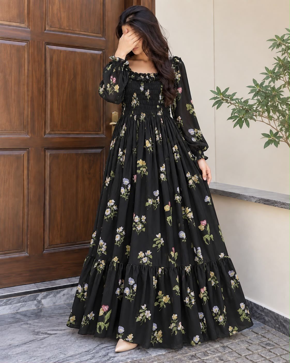 A woman wearing a long black floral patterned maxi dress standing by a wooden door.
