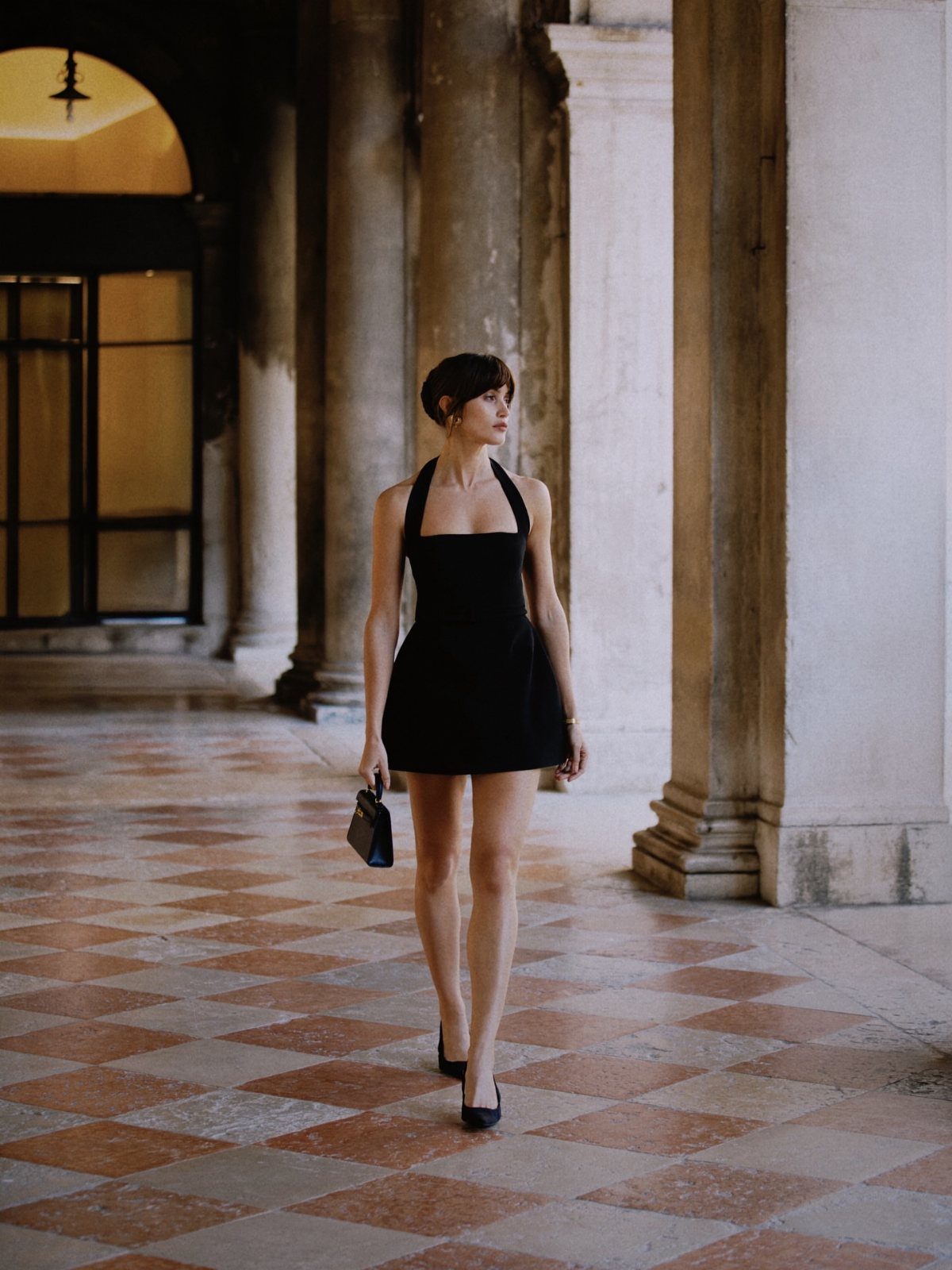 A woman in a black halter mini dress walking through a columned hallway.