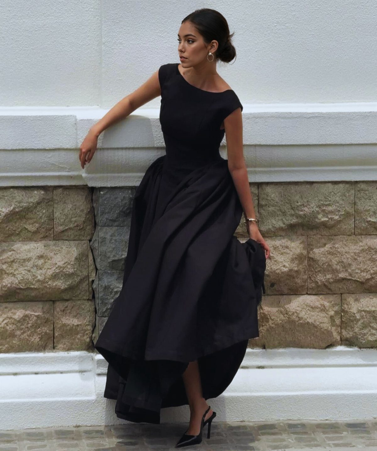 A woman in a formal black high-low dress posing against a stone and white wall.