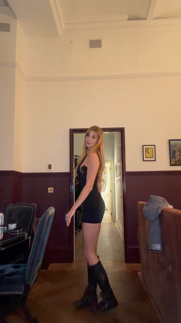 A blonde woman posing in a short, tight black dress and black boots indoors.