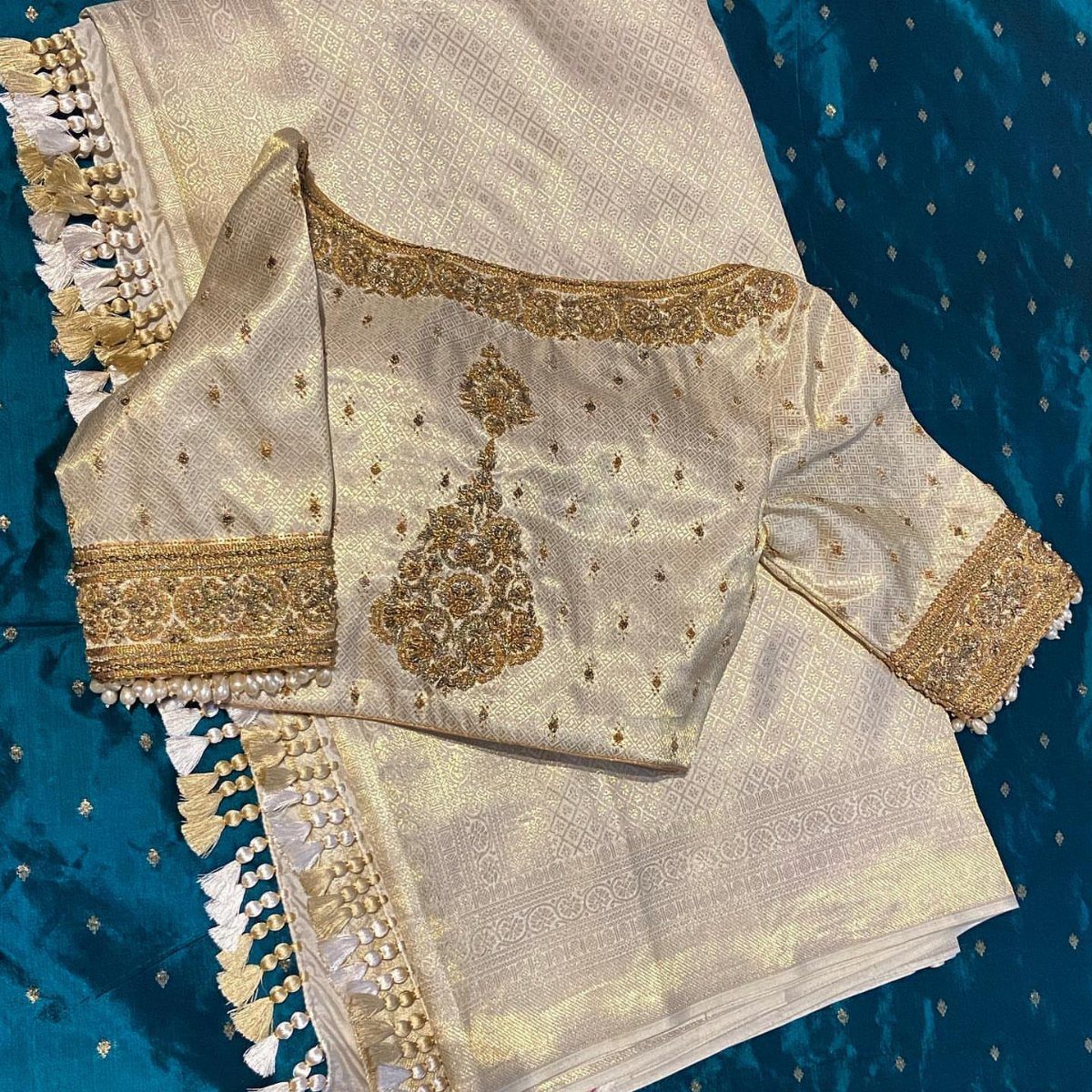 Cream silk blouse featuring intricate gold zari embroidery and patterns laid on matching fabric.