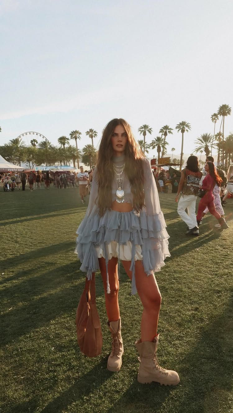 A woman wearing a ruffled blue boho outfit and tan boots standing in a grassy festival field.
