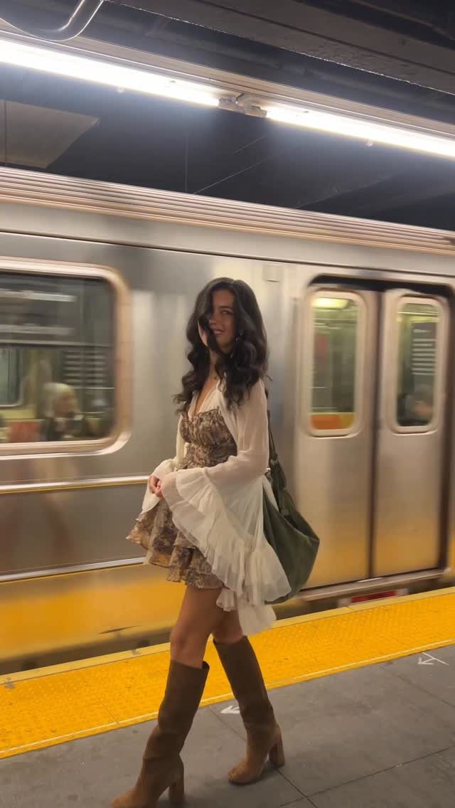 A woman wearing a boho style dress, white cardigan, and brown boots walking in a subway station.