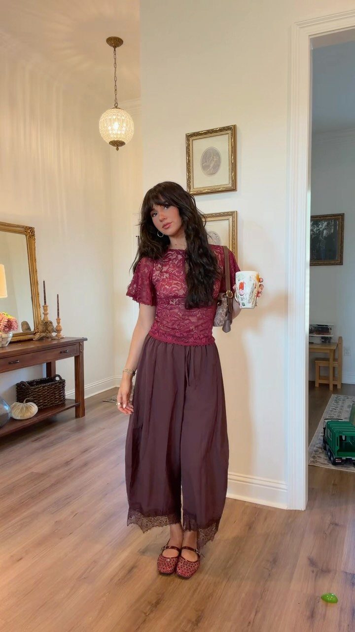A woman wearing a maroon lace top and wide-leg brown trousers standing in a room.