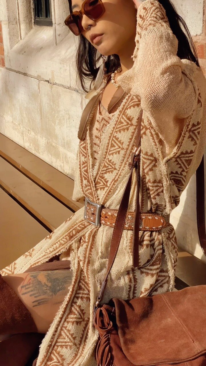 Person in a patterned cream knit dress and brown leather belt sitting in sunlight.