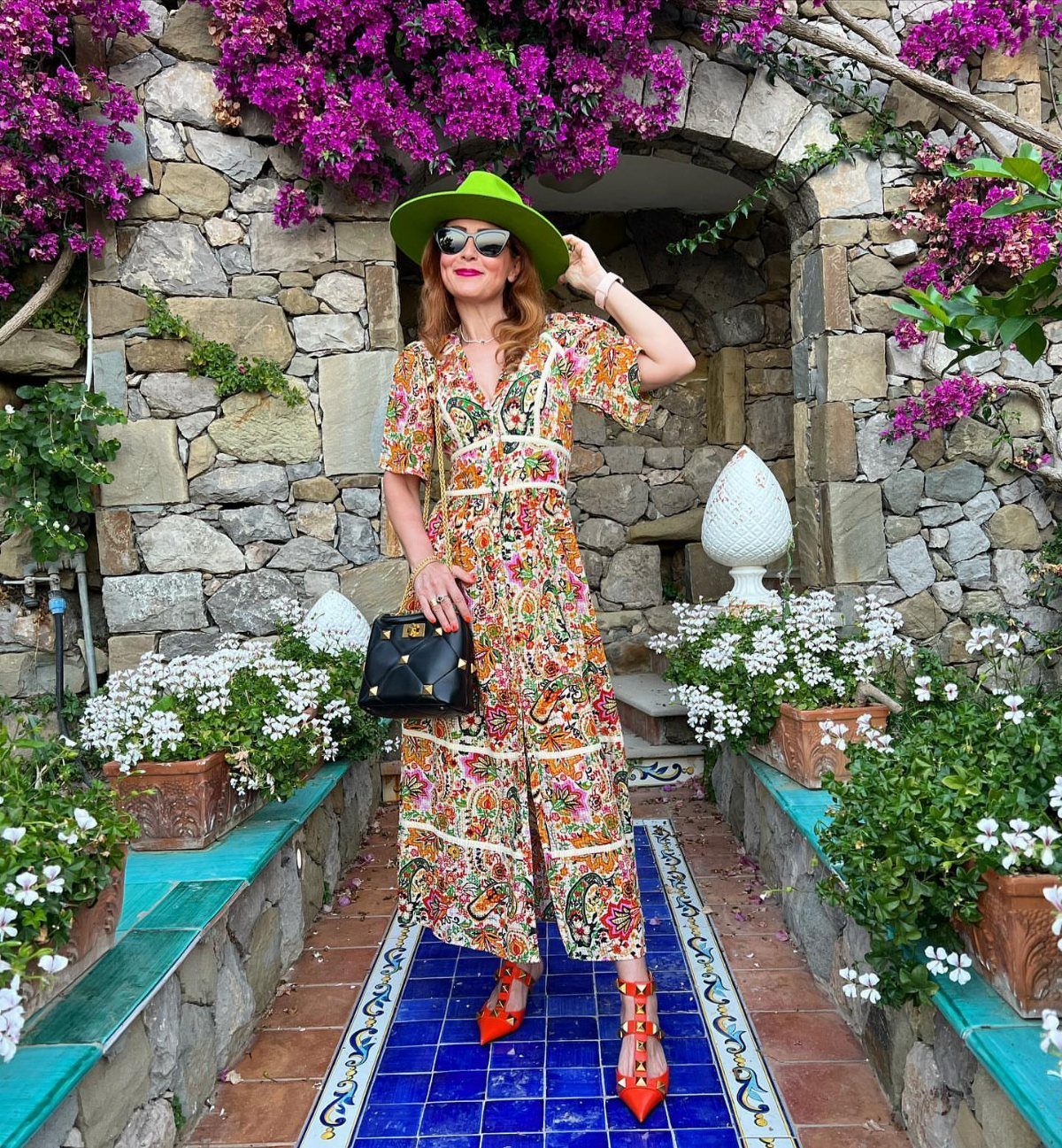Woman in a colorful floral maxi dress and green hat standing in a stone garden.