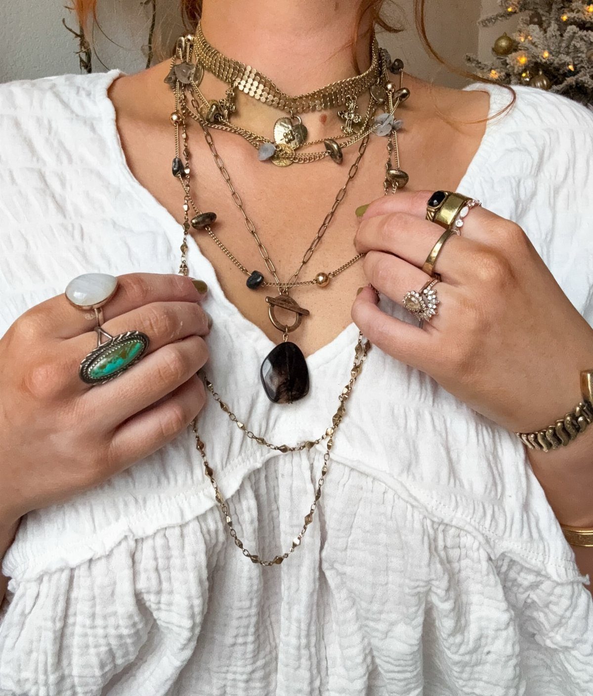 Multiple layered gold necklaces and large statement rings worn over a white textured blouse.