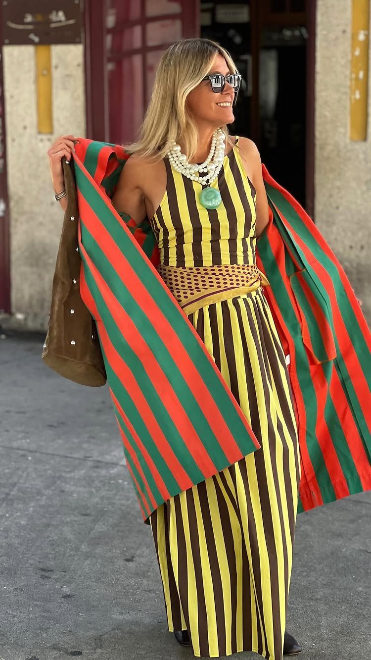 Woman in a striped jumpsuit and beaded necklace holding a colorful striped fabric outdoors.