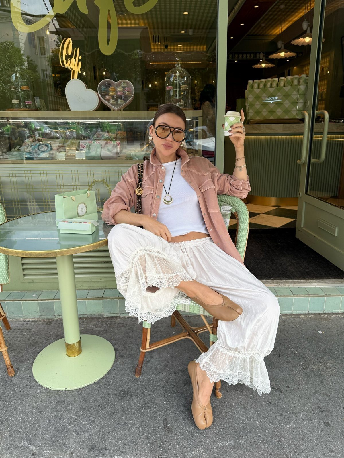 Woman in white lace trousers and pink jacket sitting at an outdoor cafe table.