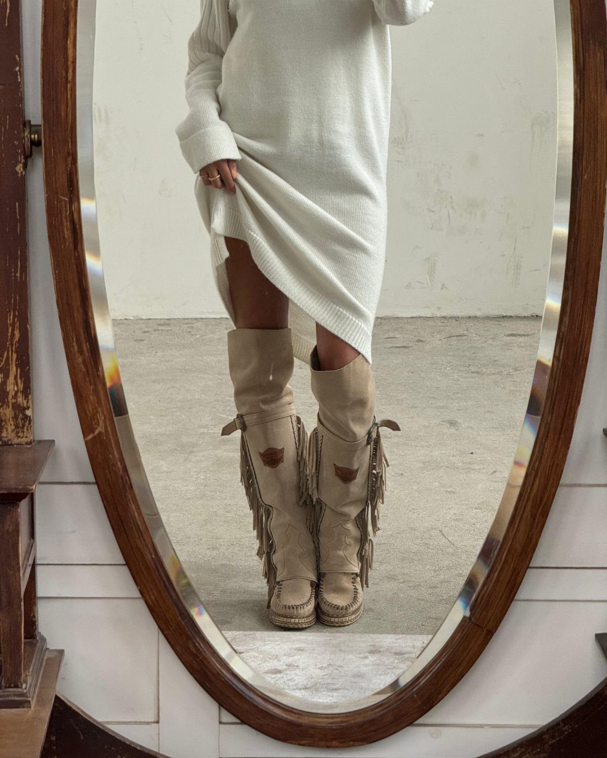 Tan suede boots with long fringe detailing worn with a white knit dress.