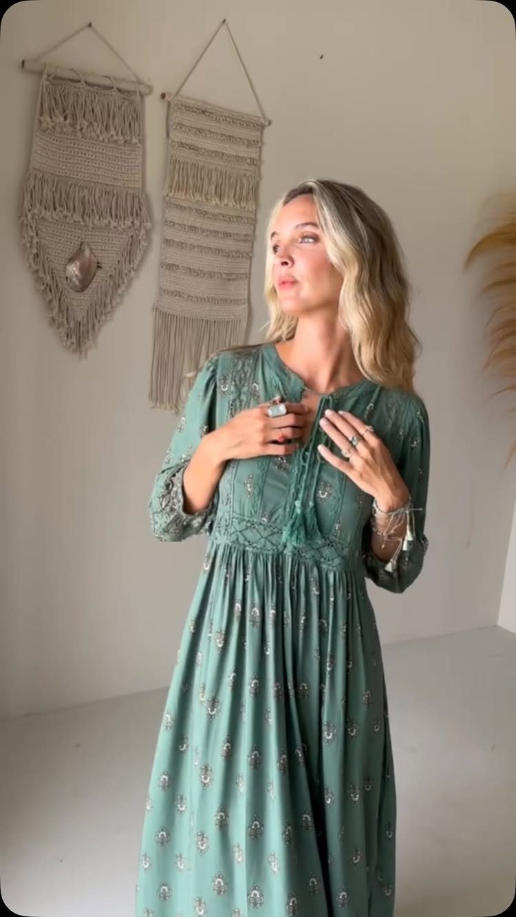 A blonde woman posing in a long, sage green patterned maxi dress with tassel details.
