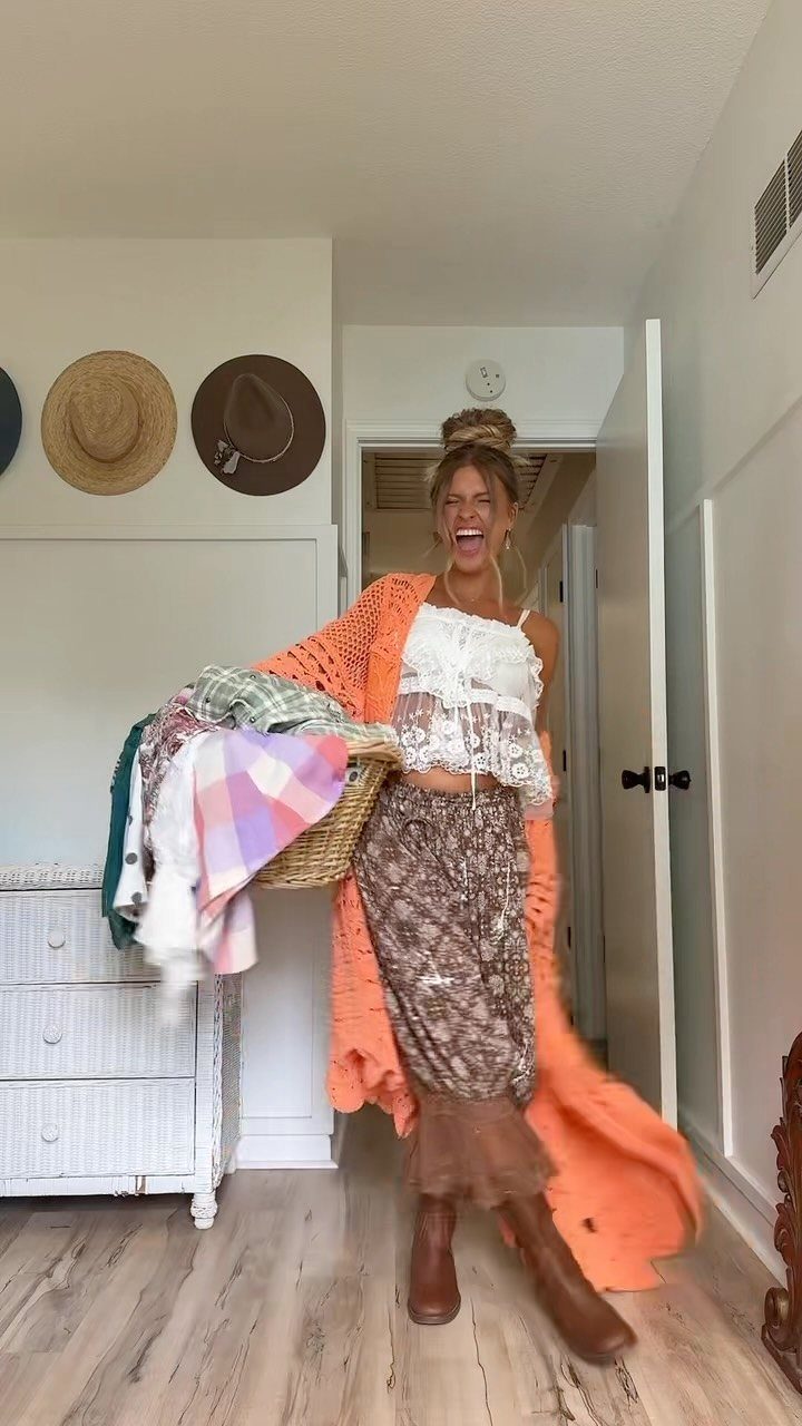 A laughing woman wearing a crochet top, patterned skirt, and orange cardigan holding a laundry basket.