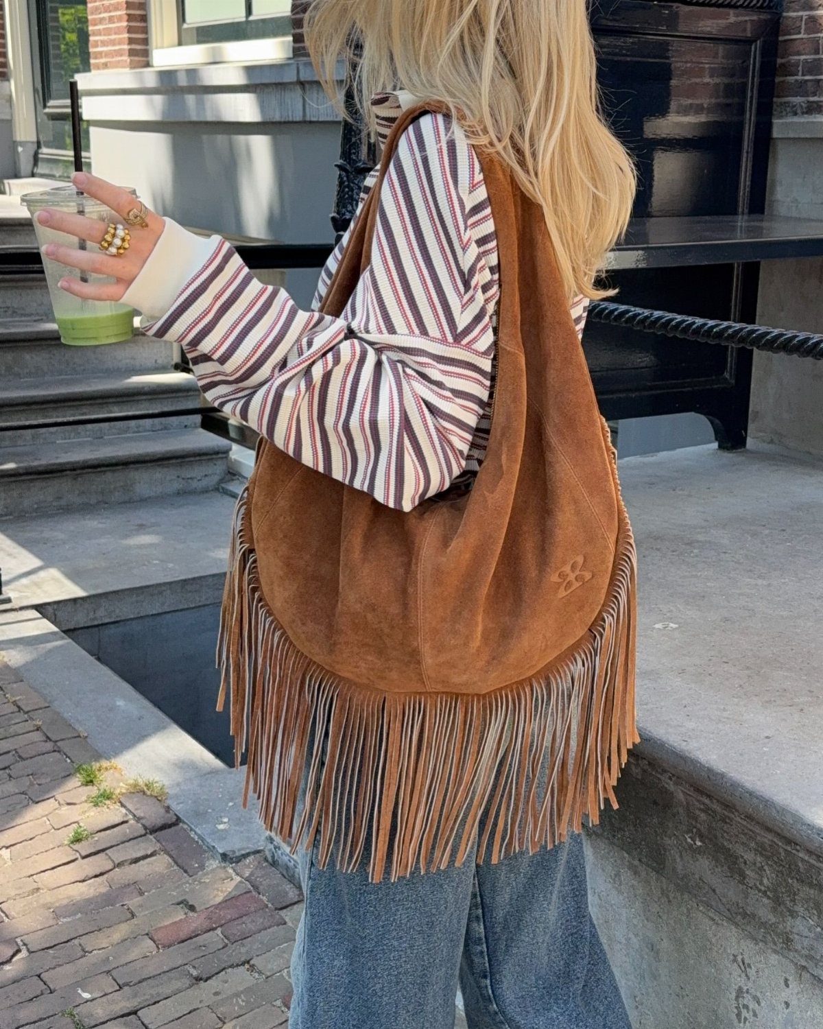 A woman carrying a brown suede fringe bag over her shoulder while holding a green drink.
