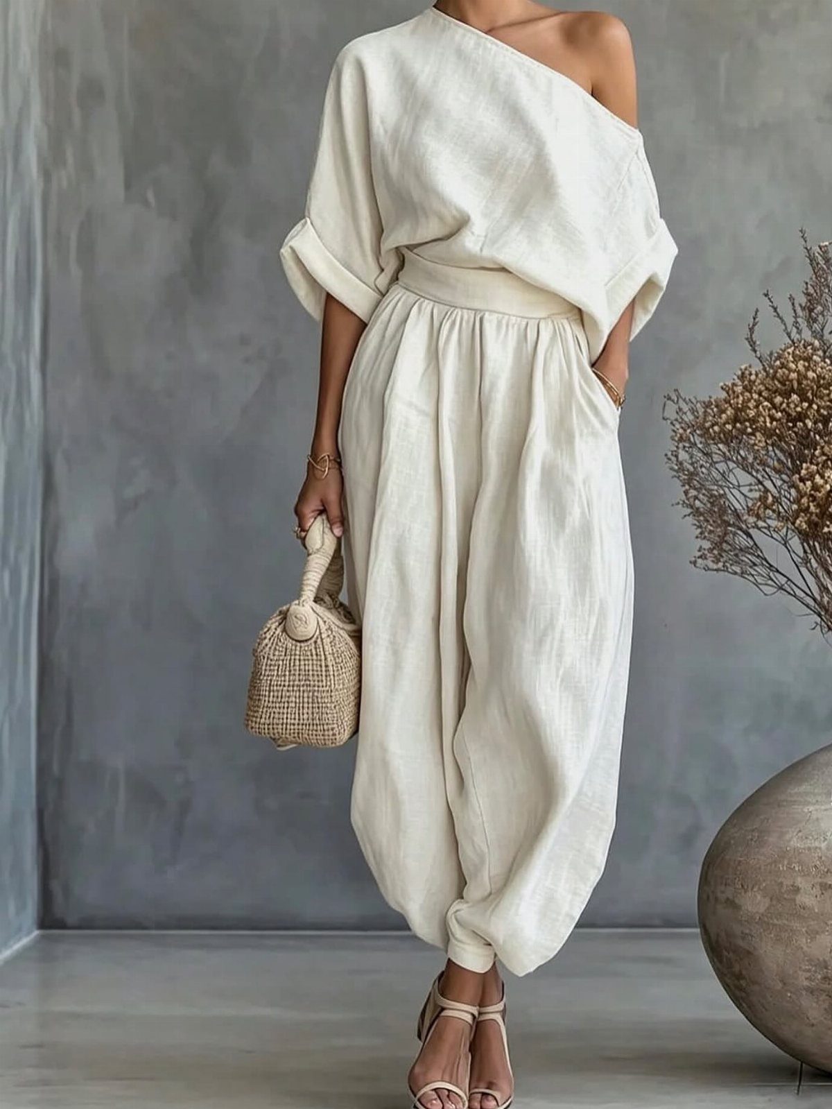 Woman wearing cream linen trousers and a matching one-shoulder top holding a straw bag.