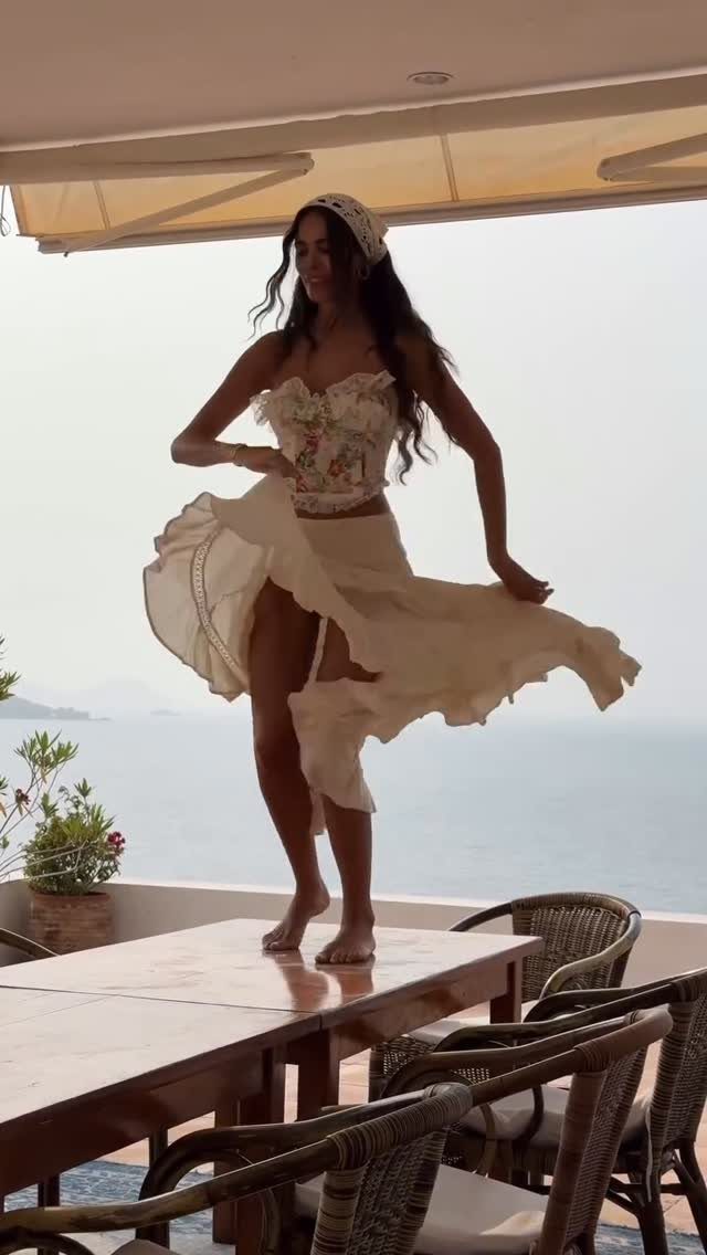 Woman in a floral corset and flowing cream maxi skirt posing on a table by the sea.