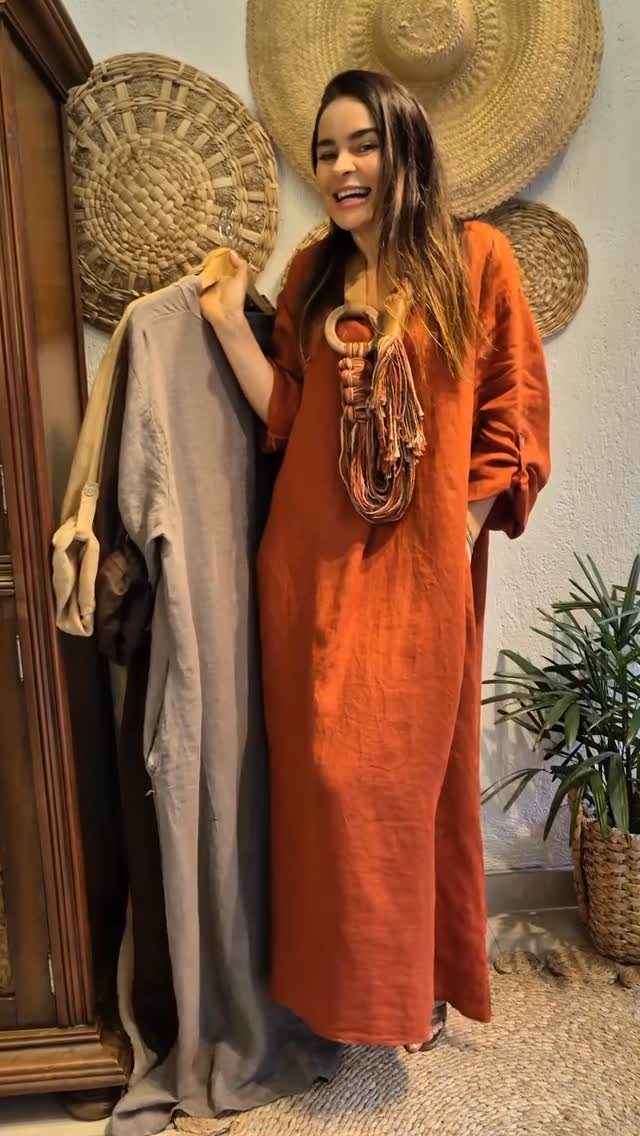 A woman in a rust-colored linen dress standing next to hanging grey clothing and woven hats.