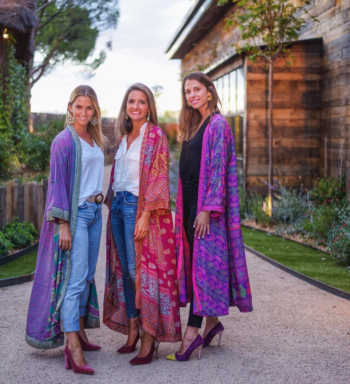 Three women posing outdoors wearing long, vibrant, patterned bohemian kimonos over casual clothing.
