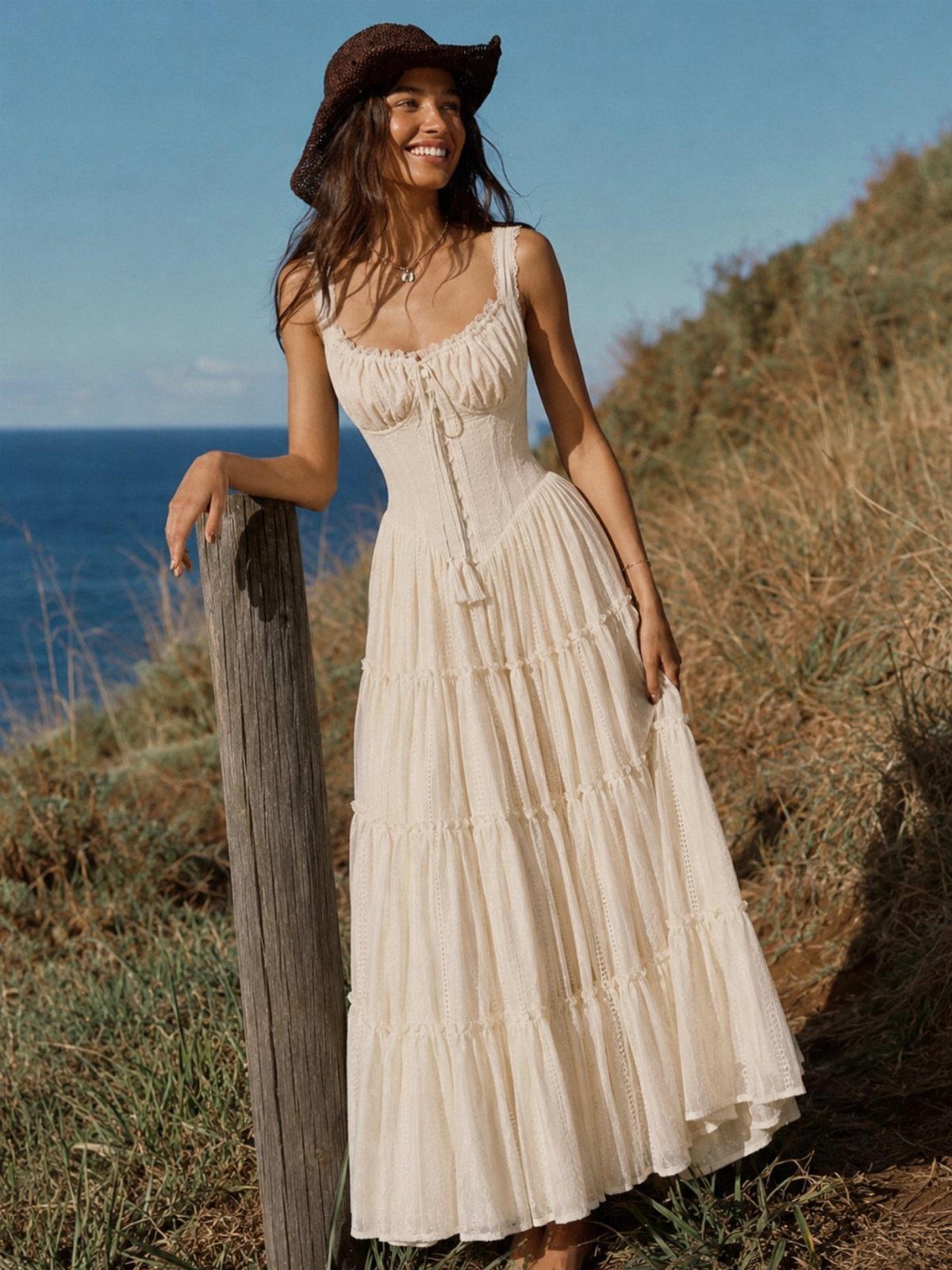 A smiling woman in a cream tiered lace maxi dress leaning against a wooden post by the ocean.