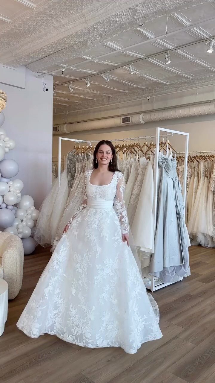 A woman smiling while wearing a white lace wedding dress with a square neckline.