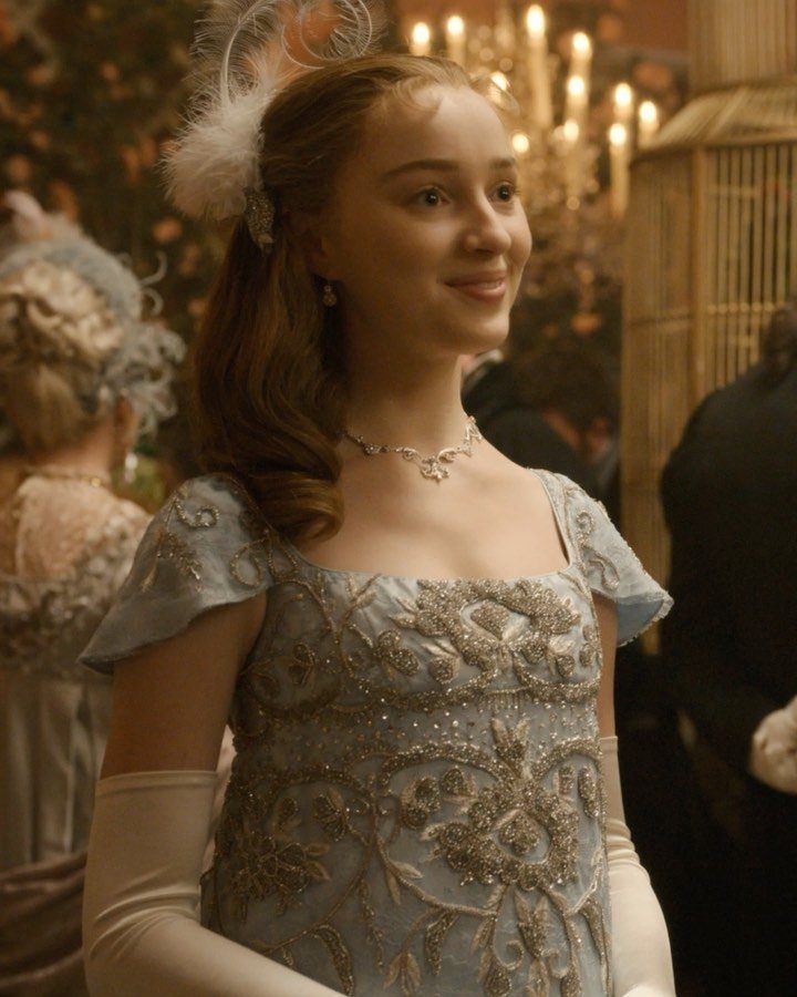 A smiling young woman wearing an embroidered light blue Regency-style dress and white gloves.