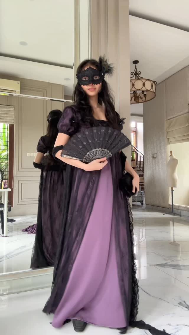 A woman in a purple lace gown and black masquerade mask holding a fan.
