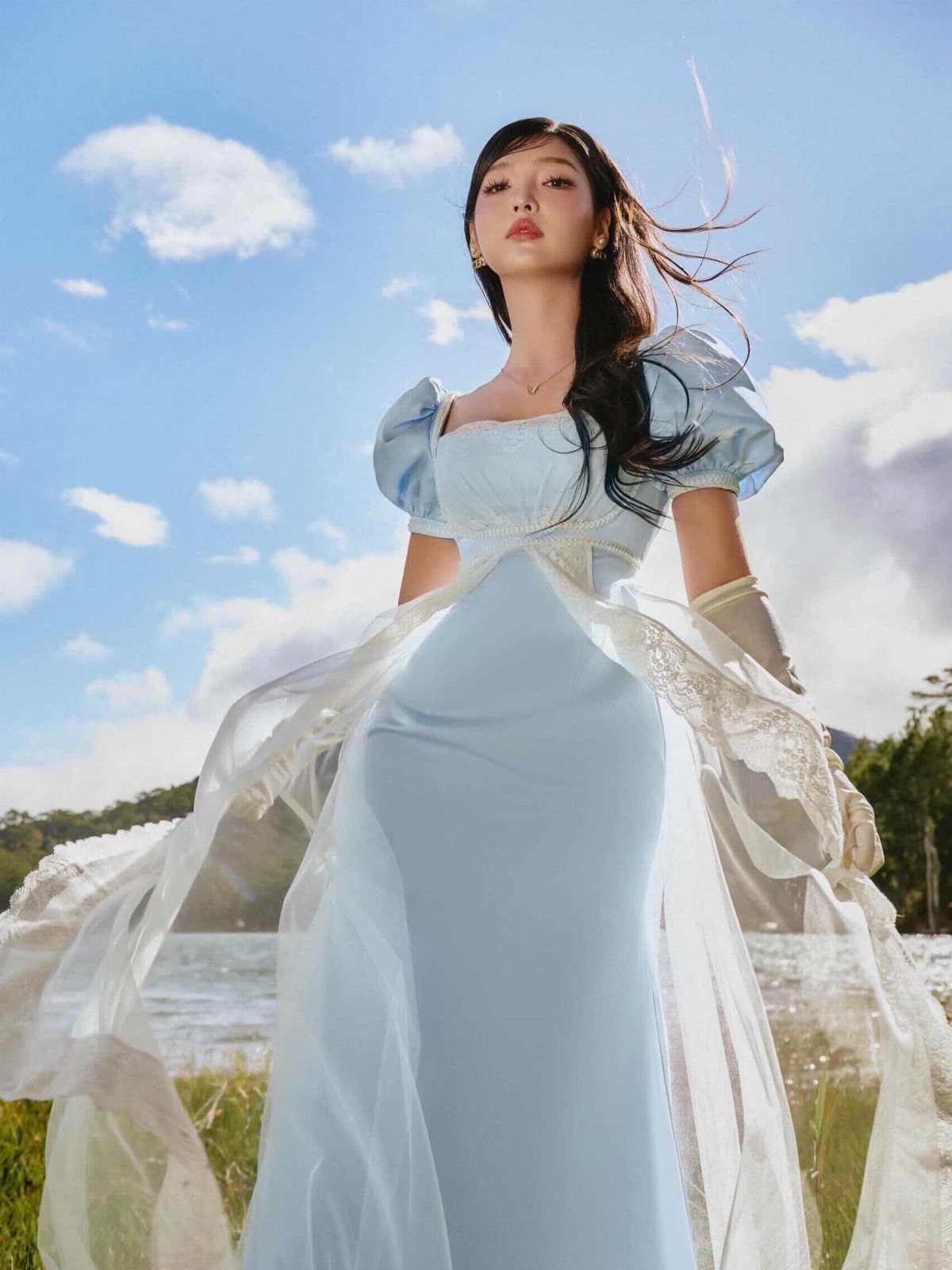 A woman wearing a light blue empire waist gown with flowing sheer fabric layers outdoors.