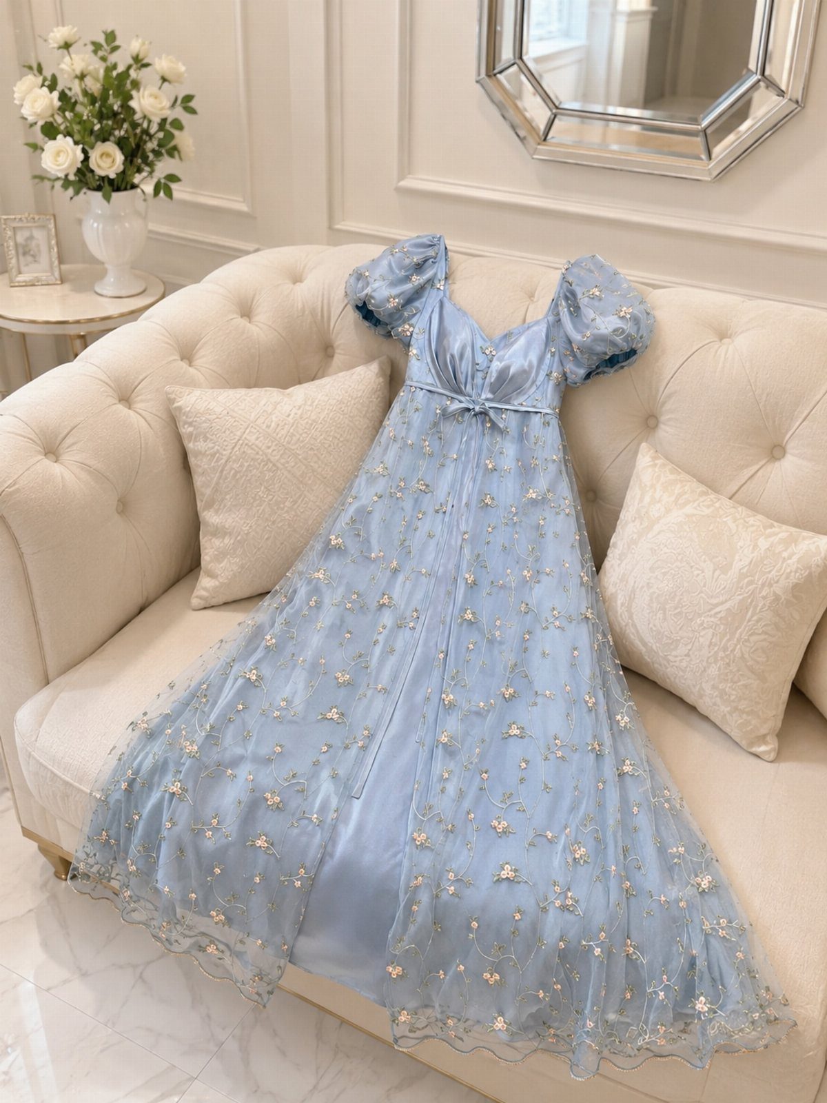 A light blue empire waist gown with puffed sleeves and floral embroidery resting on a white sofa.