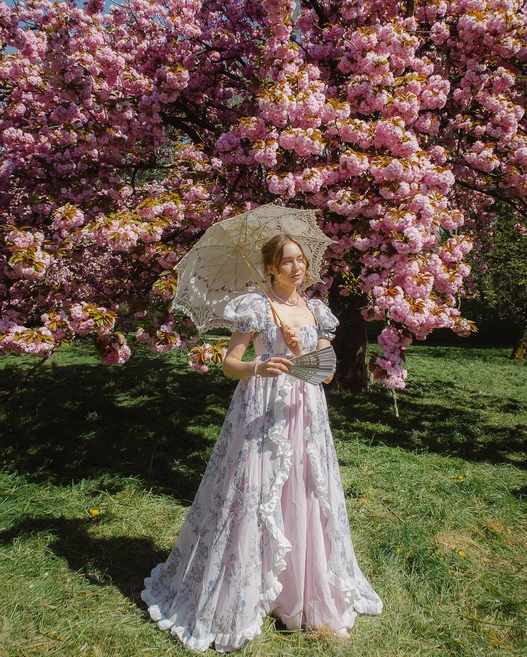 A woman wearing a floral empire-waist gown holds a lace parasol under a blooming pink tree.