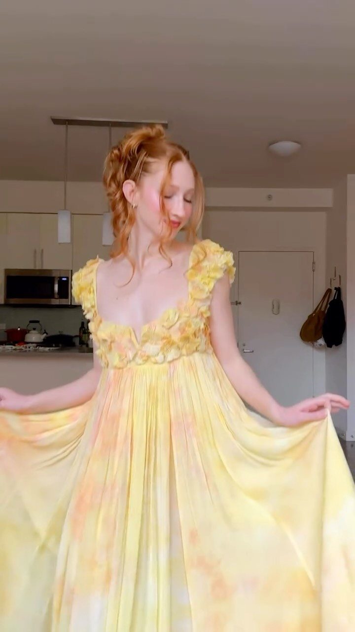A woman with red hair posing in a flowing yellow empire-waist dress with floral details.