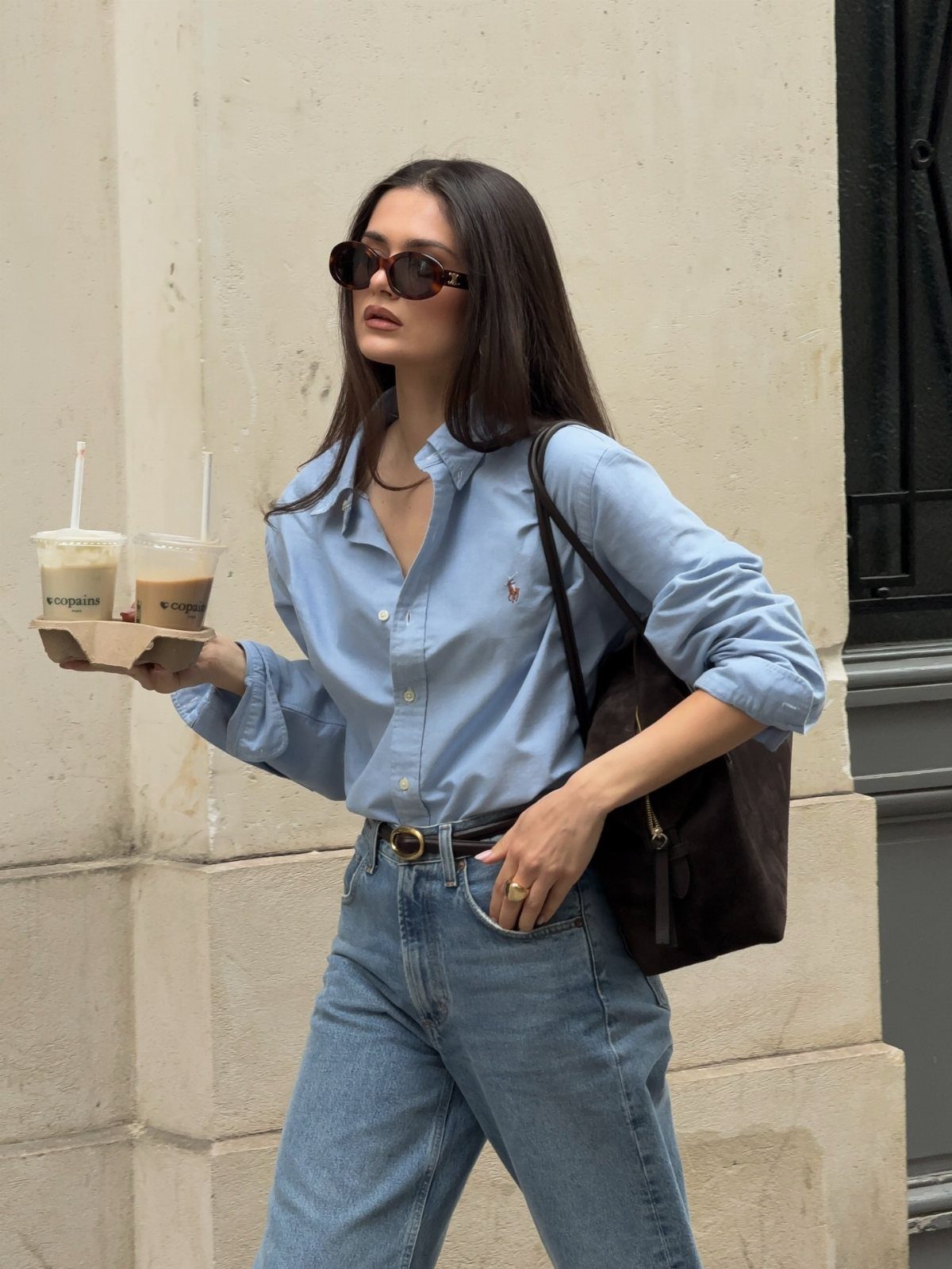 Woman in a light blue button-down shirt and blue jeans holding two iced coffees.