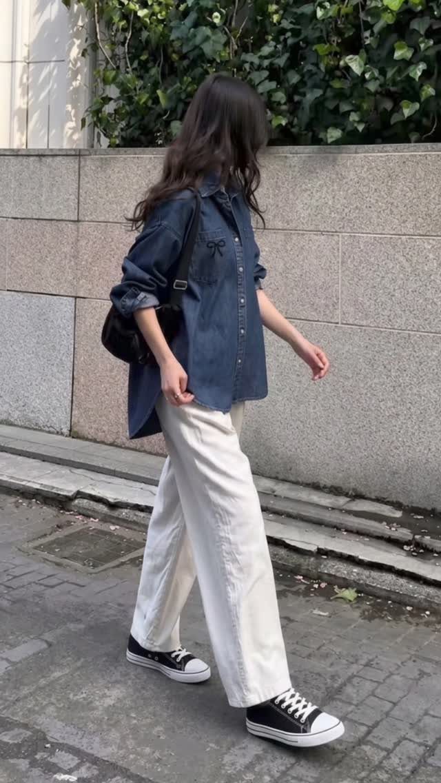 Woman walking in an oversized denim shirt and wide-leg white trousers with black sneakers.