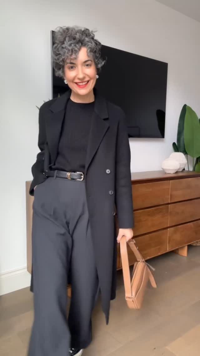 A smiling woman with curly grey hair wearing a monochromatic black outfit and holding a tan handbag.