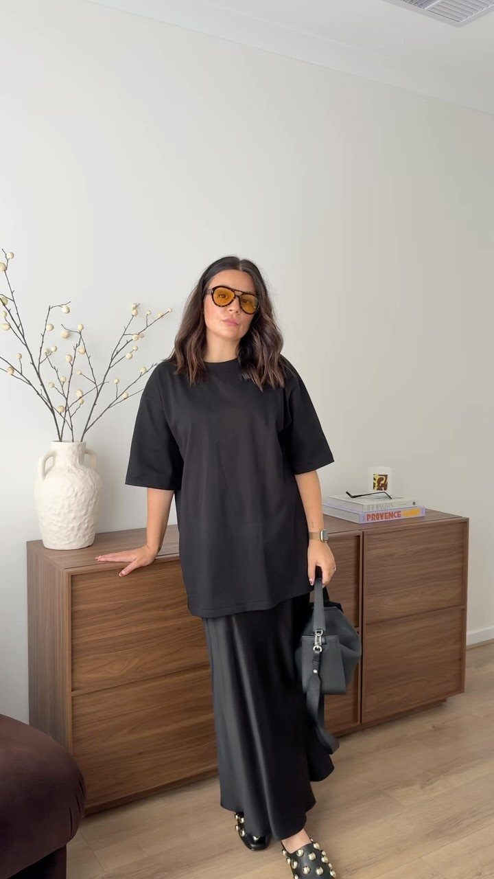 Woman in an oversized black t-shirt and long satin skirt standing by a wooden dresser.