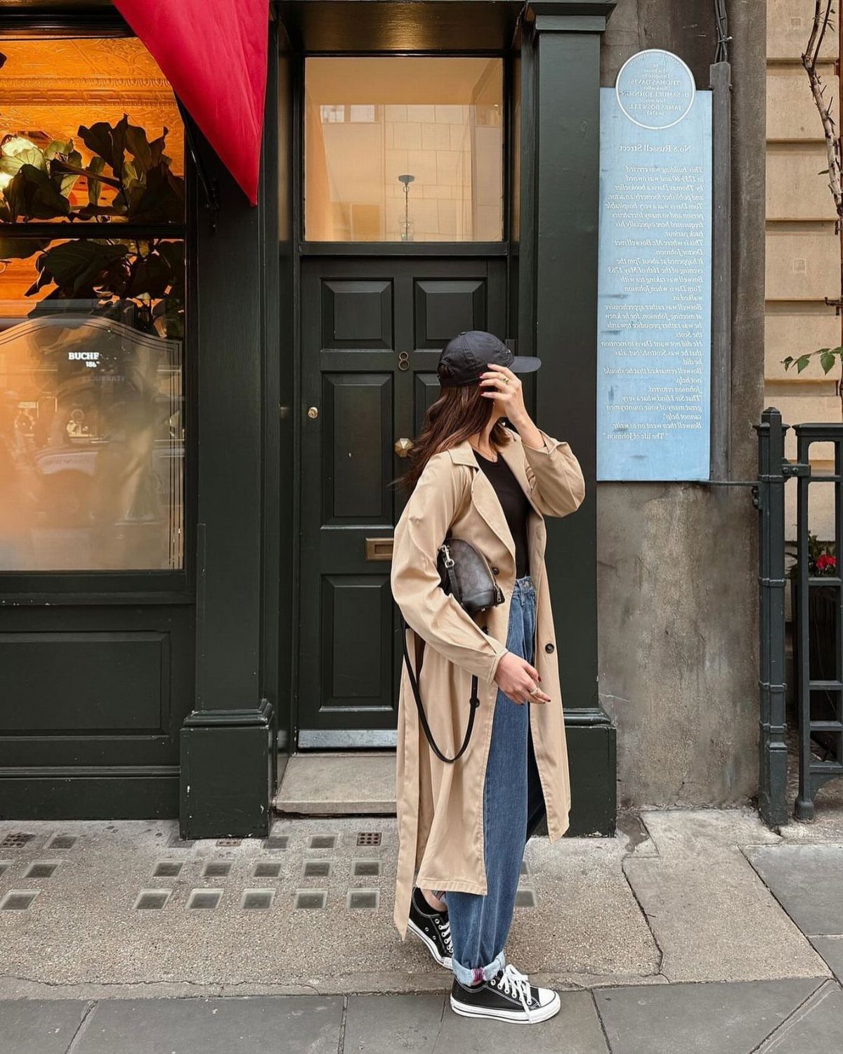 Woman wearing a tan trench coat, black top, and blue denim capris standing on a city street.