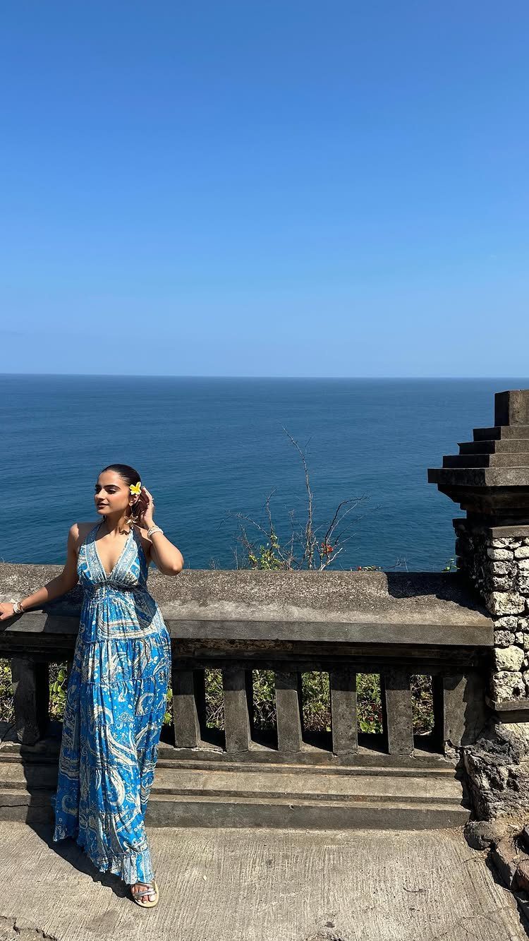 A woman wearing a blue patterned boho maxi dress posing by a stone railing overlooking the ocean.