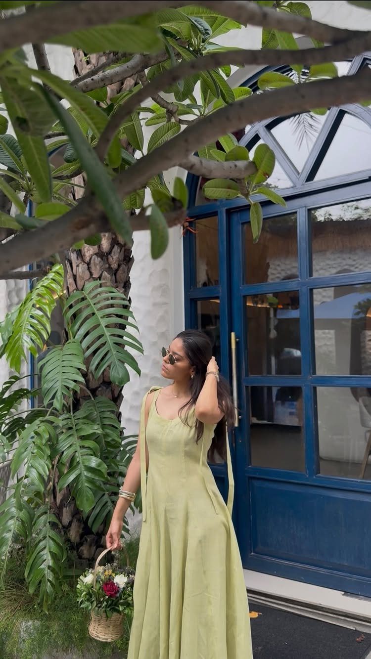 A woman in a long light green linen dress stands by a blue door and tropical greenery.