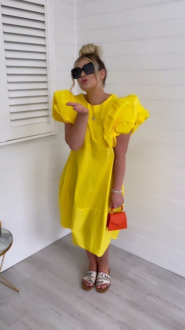 A woman in a bright yellow puff sleeve midi dress posing with an orange handbag.