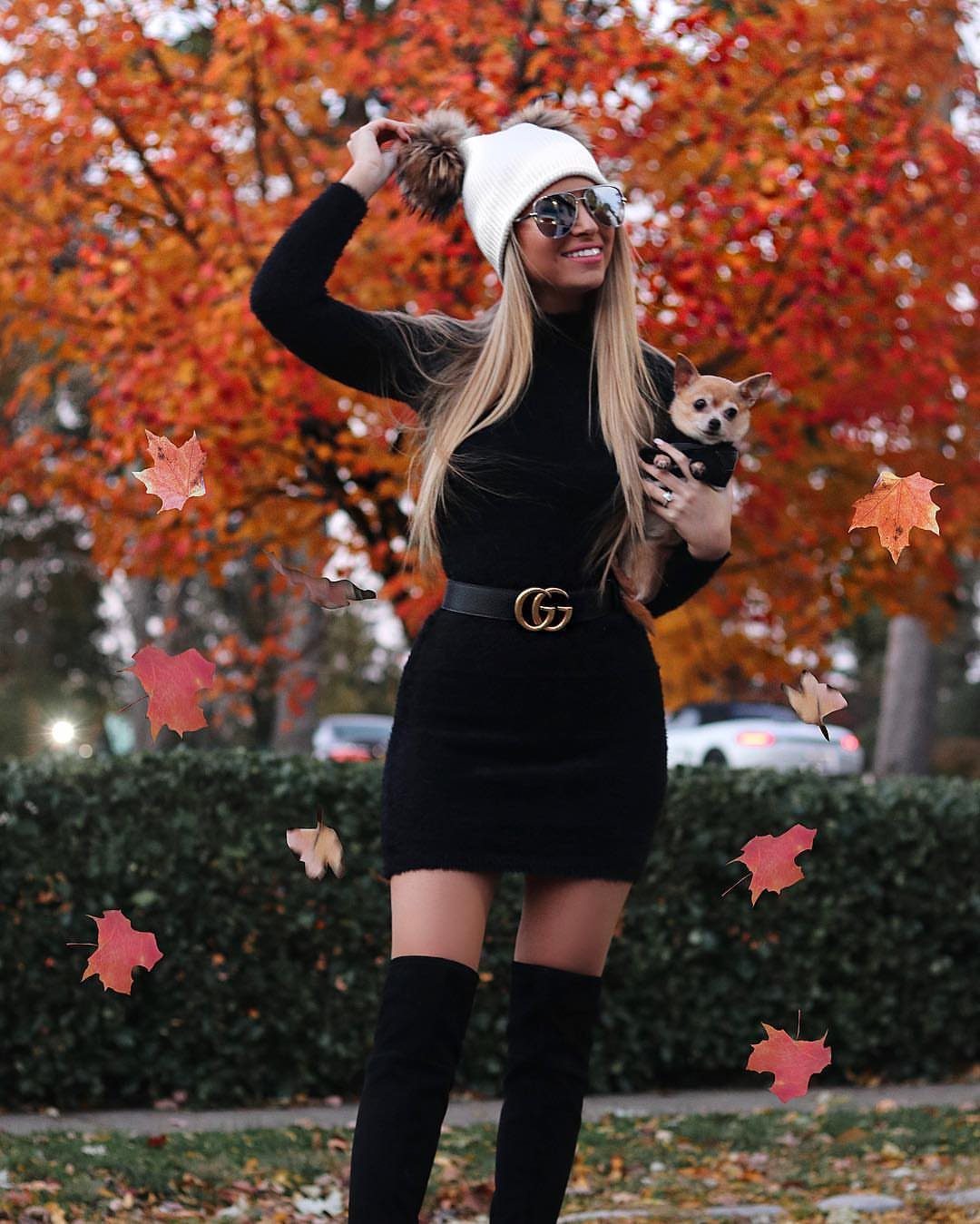 Woman in a black sweater dress and white beanie holding a small dog outdoors.